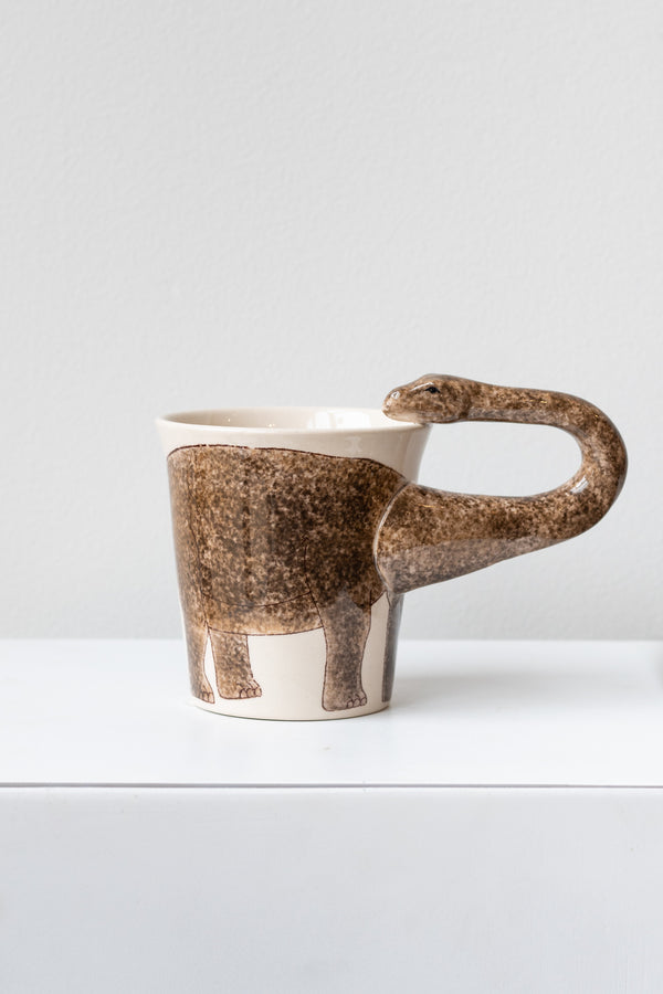 Brontosaurus Mug by Sea Island Imports sits on a white surface in a white room