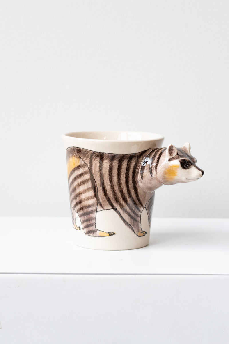 Raccoon Mug by Sea Island Imports sits on a white surface in a white room