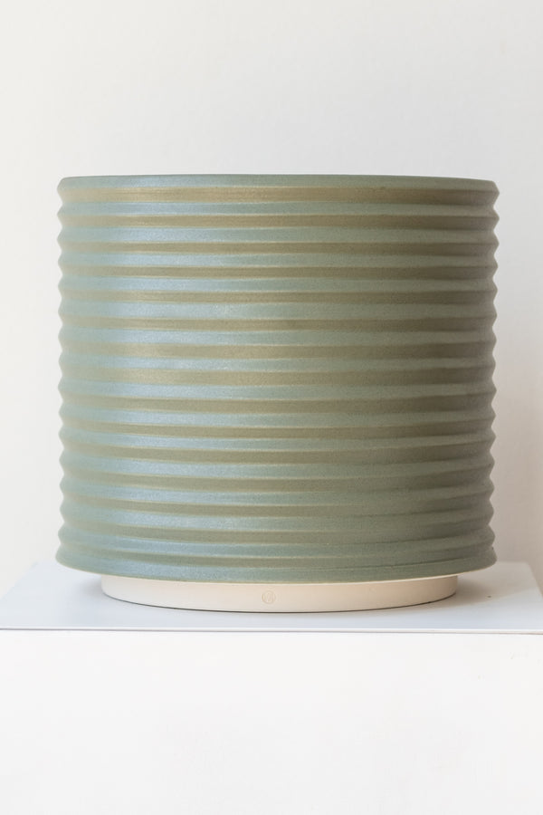 A large grey-green planter sits on a white surface in a white room. The planter is cylindrical and ribbed. It sits on an unglazed ceramic drainage tray. The planter is empty. It is photographed straight on.
