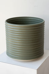 A large grey-green planter sits on a white surface in a white room. The planter is cylindrical and ribbed. It sits on an unglazed ceramic drainage tray. The planter is empty. It is photographed closer and at an angle.