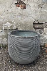 A frontal view of Cody Pot horizontal ridged dark grey medium against a concrete backdrop