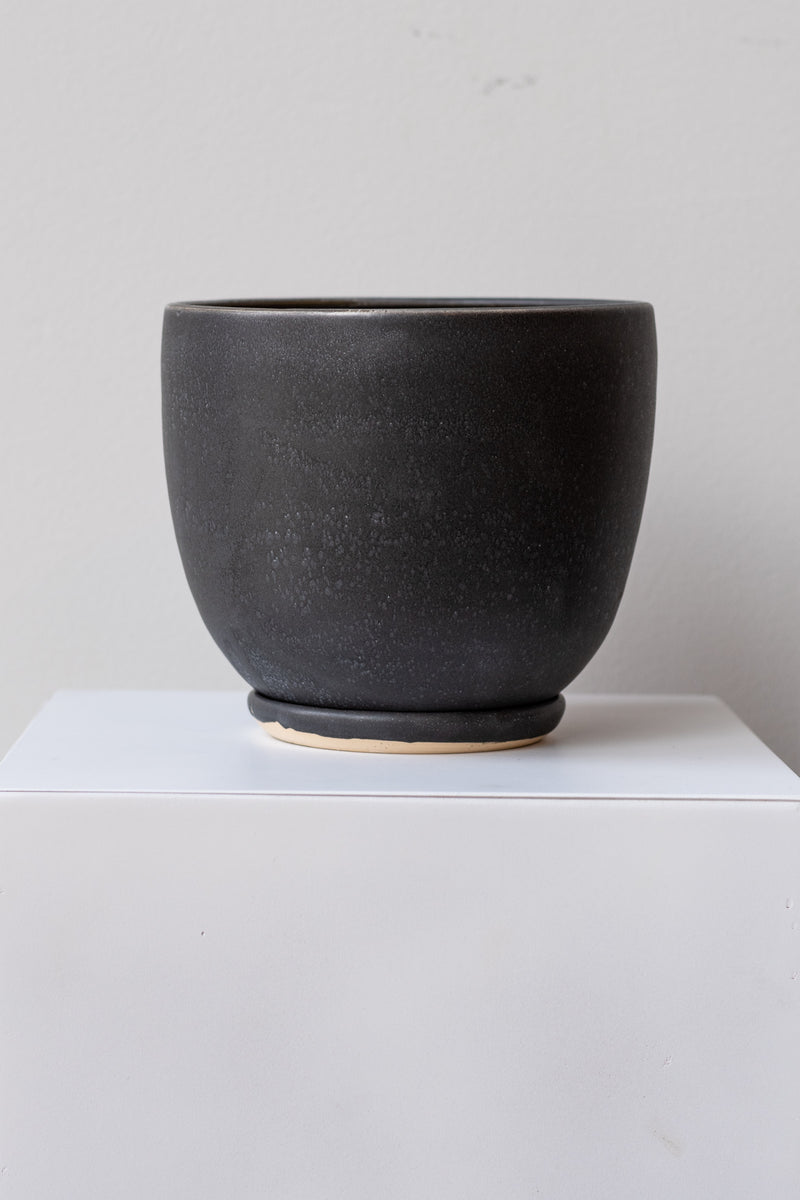 One large ceramic planter sits on a white surface in a white room. The planter is glazed black with white speckles. It sits on a small drainage tray. The planter is empty. It is photographed straight on.