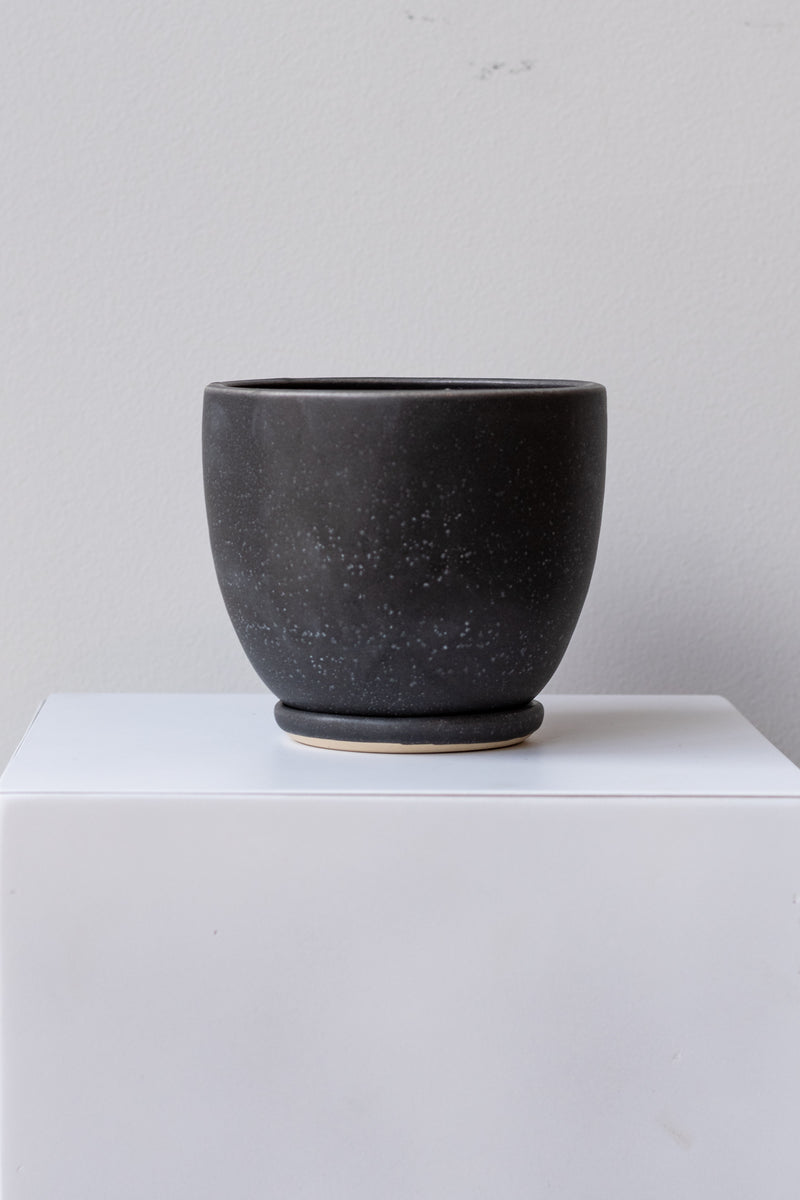 One small ceramic planter sits on a white surface in a white room. The planter is glazed black with white speckles. It sits on a small drainage tray. The planter is empty. It is photographed straight on.