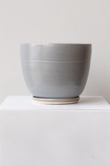 One large ceramic planter sits on a white surface in a white room. The planter is glazed grey. It sits on a small drainage tray. The planter is empty. It is photographed straight on.