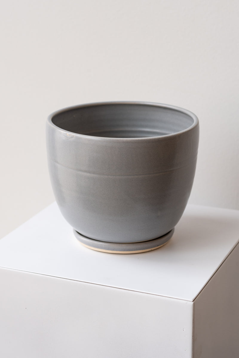 One large ceramic planter sits on a white surface in a white room. The planter is glazed grey. It sits on a small drainage tray. The planter is empty. It is photographed closer and at an angle.
