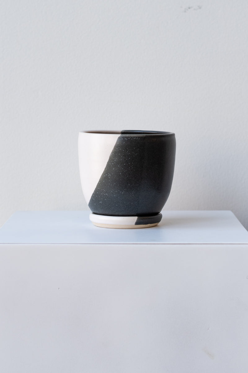 One small ceramic planter sits on a white surface in a white room. The planter is glazed half black and half white. It sits on a small drainage tray which is also half black and half white. The planter is empty. It is photographed straight on.