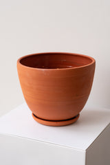 One large terra cotta planter sits on a white surface in a white room. The planter sits on a small round drainage tray. The planter is empty. It is photographed closer and at an angle. The inside of the planter is glazed.