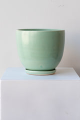 One large ceramic planter sits on a white surface in a white room. The planter is glazed light blue-green. It sits on a small drainage tray. The planter is empty. It is photographed straight on.
