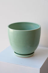 One large ceramic planter sits on a white surface in a white room. The planter is glazed light blue-green. It sits on a small drainage tray. The planter is empty. It is photographed closer and at an angle.