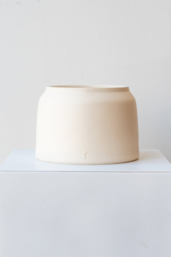 One cream colored stoneware planter sits on a white surface in a white room. The planter is round and squat, with a small logo imprinted in the clay on the bottom. It is photographed straight on.