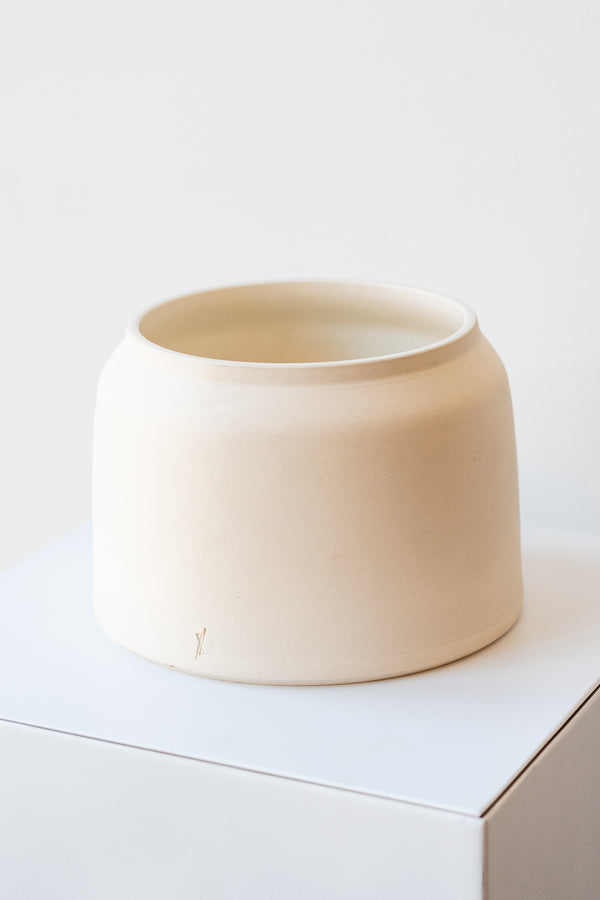 One cream colored stoneware planter sits on a white surface in a white room. The planter is round and squat, with a small logo imprinted in the clay on the bottom. It is photographed at an angle and slightly closer to show the clay detail. 