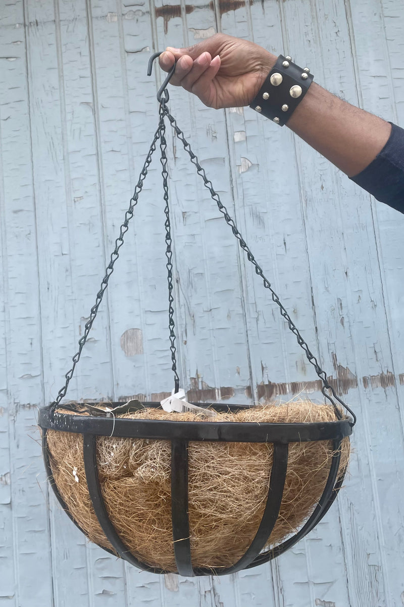 A hand holds Forge Hanging Basket w/ Coco Liner 14" against wooden backdrop