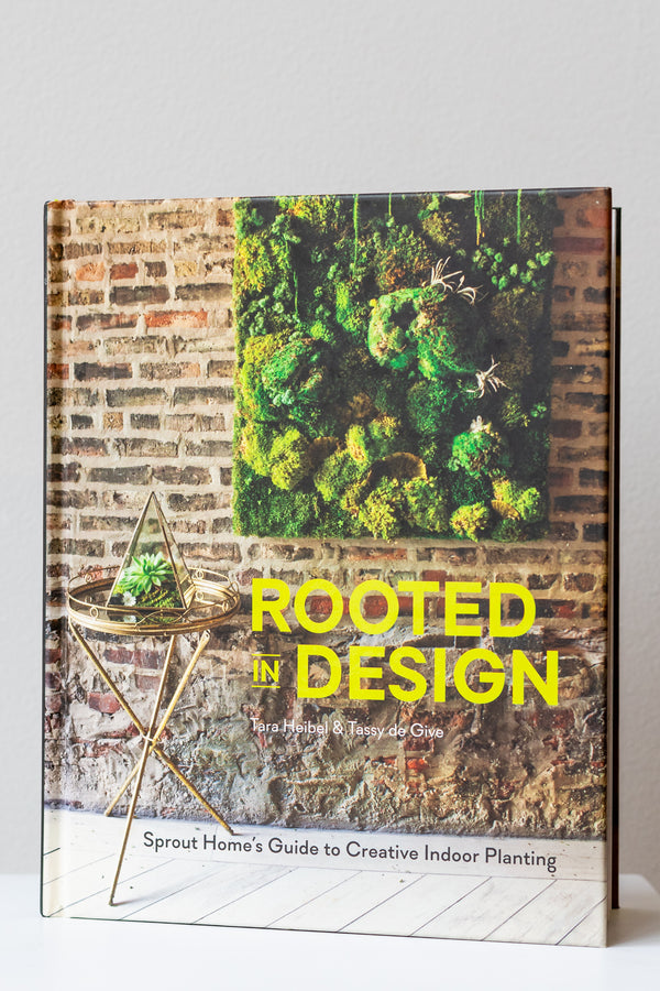 The book "Rooted In Design" by Tara Heibel and Tassy de Give sits on a white surface in a white room. The cover of the book has a photo of a brick wall with a large moss wall hanging, as well as a small gold table with a pyramid-shaped terrarium. It is photographed straight on.