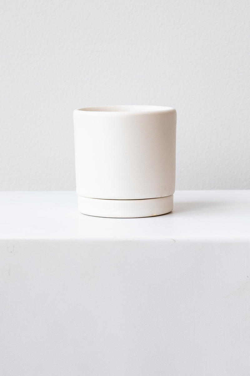 A small white ceramic planter sits on a white surface in a white room. The planter has a matching drainage tray. The planter is empty. It is photographed straight on.