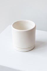 A small white ceramic planter sits on a white surface in a white room. The planter has a matching drainage tray. The planter is empty. It is photographed closer and at an angle.