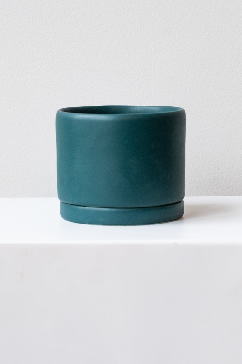 A medium dark teal ceramic planter sits on a white surface in a white room. The planter has a matching drainage tray. The planter is empty. It is photographed straight on.