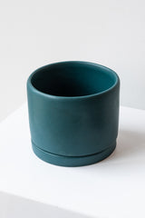 A medium dark teal ceramic planter sits on a white surface in a white room. The planter has a matching drainage tray. The planter is empty. It is photographed closer and at an angle.
