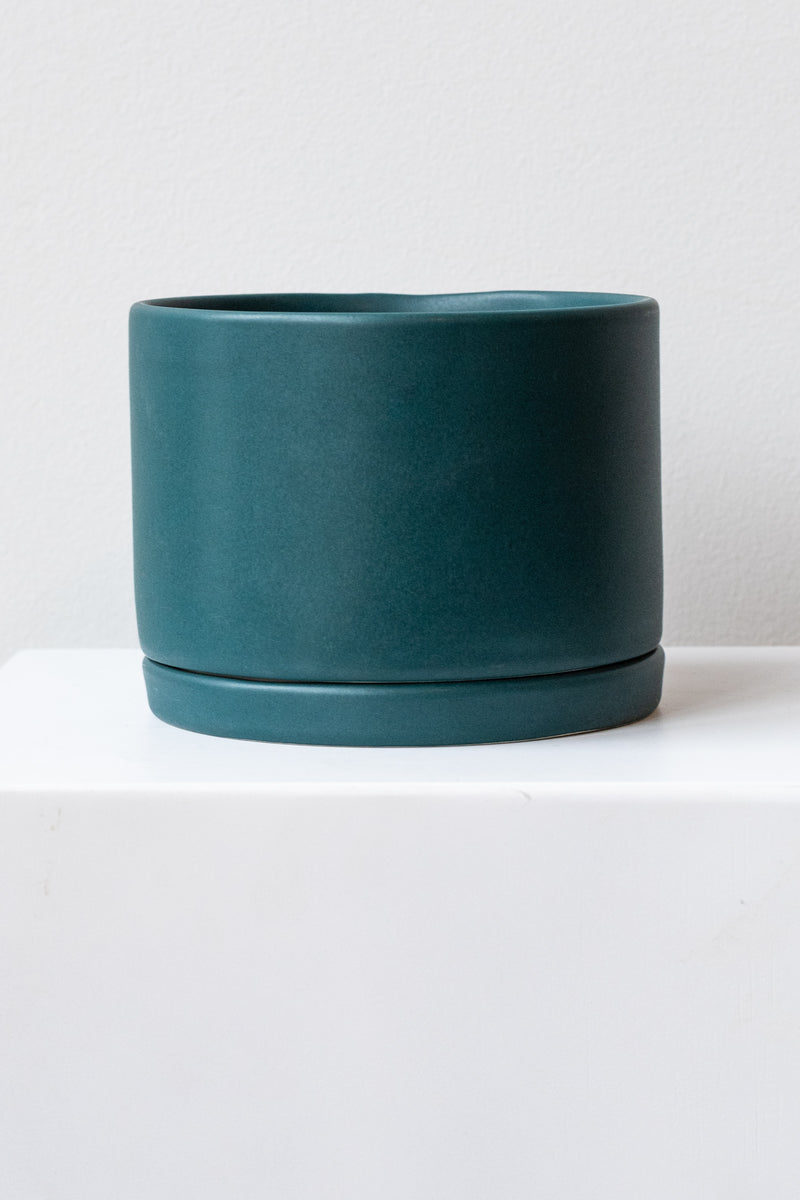 A large dark teal ceramic planter sits on a white surface in a white room. The planter has a matching drainage tray. The planter is empty. It is photographed straight on.