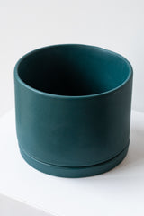 A large dark teal ceramic planter sits on a white surface in a white room. The planter has a matching drainage tray. The planter is empty. It is photographed closer and at an angle.