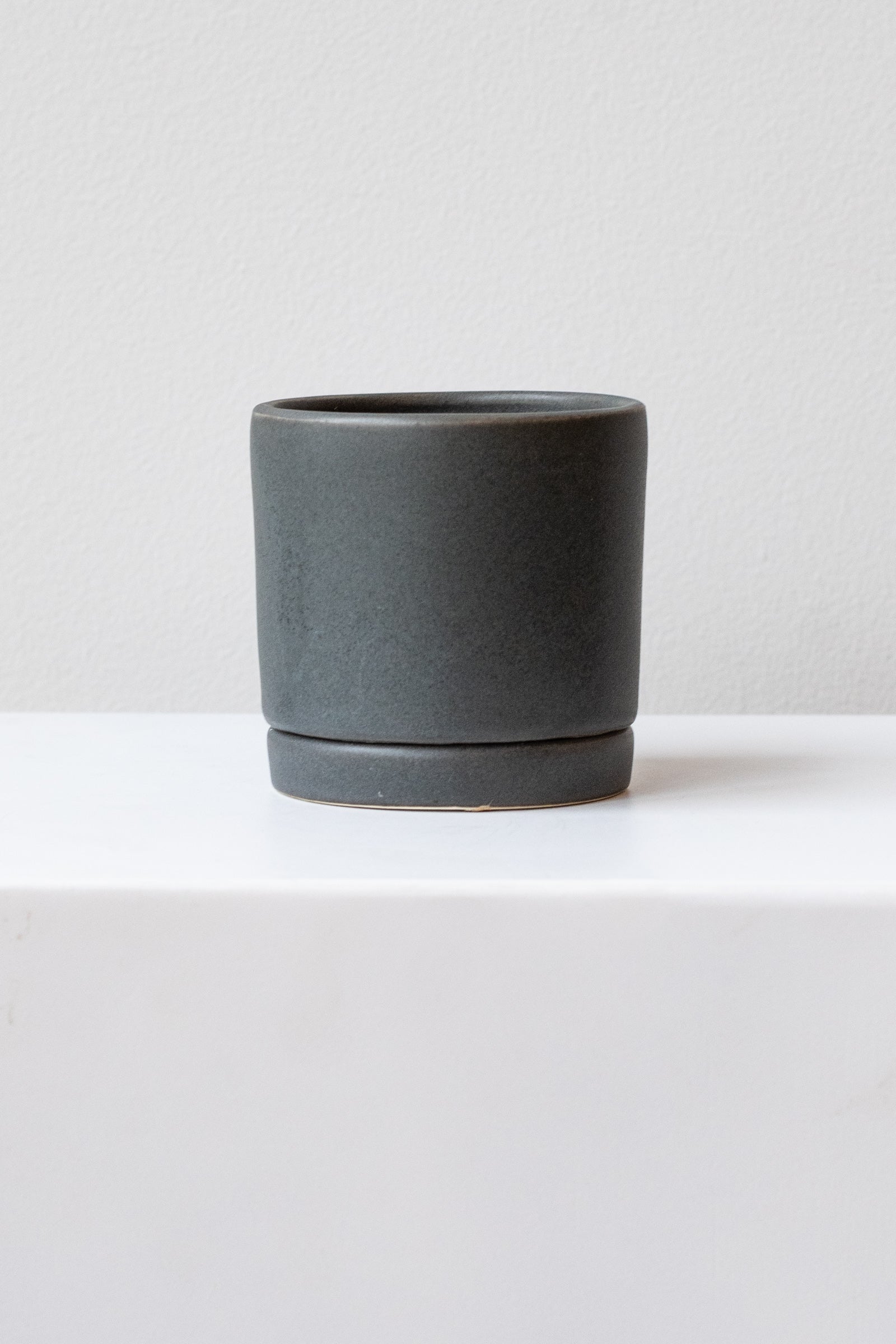 A small charcoal ceramic planter sits on a white surface in a white room. The planter has a matching drainage tray. The planter is empty. It is photographed straight on.