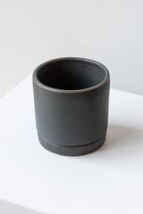 A small charcoal ceramic planter sits on a white surface in a white room. The planter has a matching drainage tray. The planter is empty. It is photographed closer and at an angle.