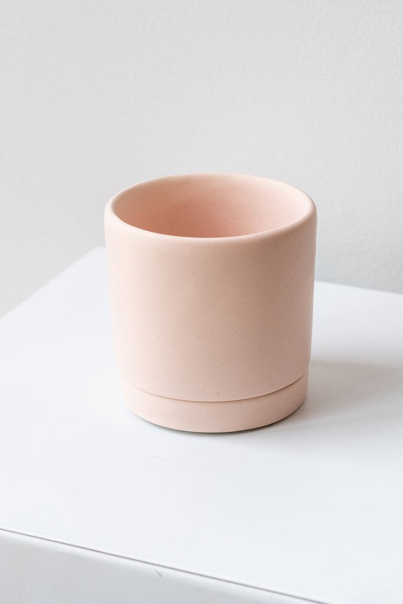 A small pink ceramic planter sits on a white surface in a white room. The planter has a matching drainage tray. The planter is empty. It is photographed closer and at an angle.