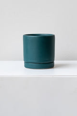 A small dark teal ceramic planter sits on a white surface in a white room. The planter has a matching drainage tray. The planter is empty. It is photographed straight on.