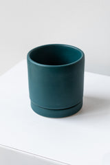 A small dark teal ceramic planter sits on a white surface in a white room. The planter has a matching drainage tray. The planter is empty. It is photographed closer and at an angle.