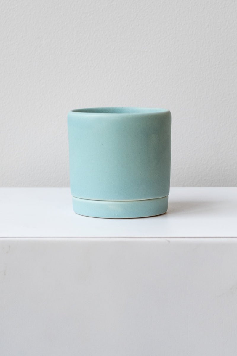 A small light blue ceramic planter sits on a white surface in a white room. The planter has a matching drainage tray. The planter is empty. It is photographed straight on.