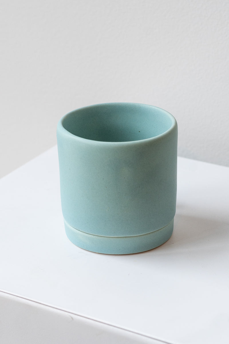 A small light blue ceramic planter sits on a white surface in a white room. The planter has a matching drainage tray. The planter is empty. It is photographed closer and at an angle.