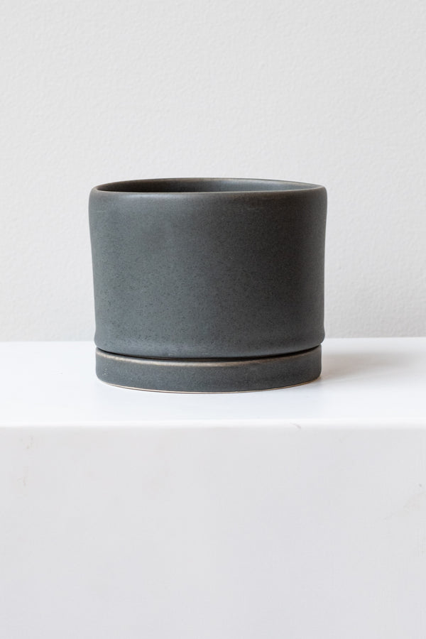 A medium charcoal ceramic planter sits on a white surface in a white room. The planter has a matching drainage tray. The planter is empty. It is photographed straight on.