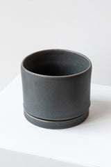 A medium charcoal ceramic planter sits on a white surface in a white room. The planter has a matching drainage tray. The planter is empty. It is photographed closer and at an angle.