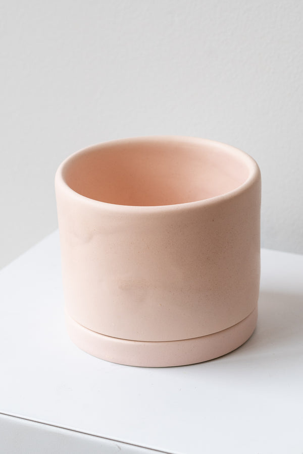 A medium pink ceramic planter sits on a white surface in a white room. The planter has a matching drainage tray. The planter is empty. It is photographed closer and at an angle.
