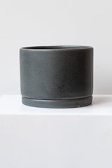 A large charcoal ceramic planter sits on a white surface in a white room. The planter has a matching drainage tray. The planter is empty. It is photographed straight on.