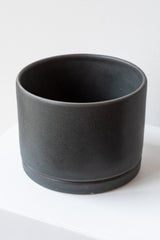 A large charcoal ceramic planter sits on a white surface in a white room. The planter has a matching drainage tray. The planter is empty. It is photographed closer and at an angle.
