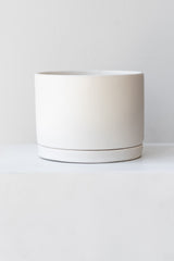 A large white ceramic planter sits on a white surface in a white room. The planter has a matching drainage tray. The planter is empty. It is photographed straight on.