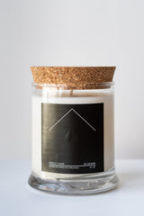 A white candle in a glass jar sits on a white surface in a white room. The candle has a cork lid and a black label with white text. The text reads: "sprout home, by: einnim, hand poured in Chicago, 10 oz"