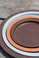 Stacked clay, copper, and granite plant saucers in front of grey wood background