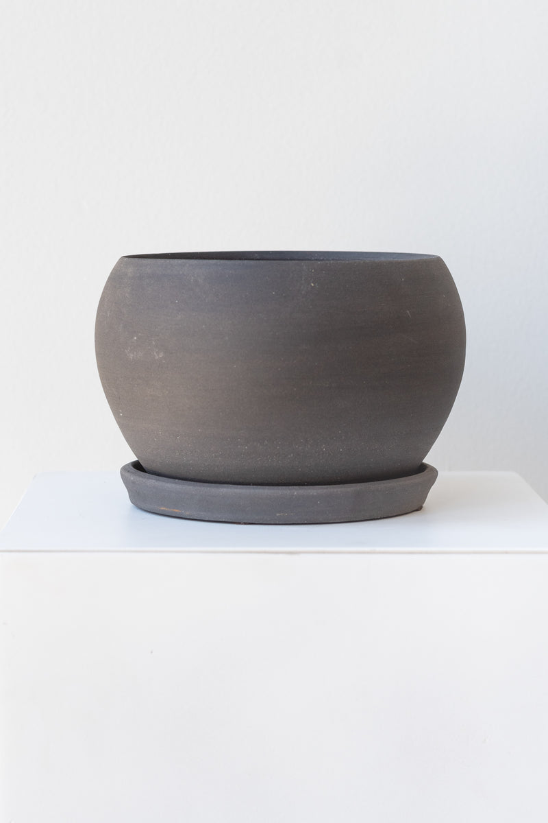 A round grey ceramic planter sits on a white surface in a white room. The planter is bubble-shaped and sits on a round drainage tray. The planter is empty. It is photographed straight on.
