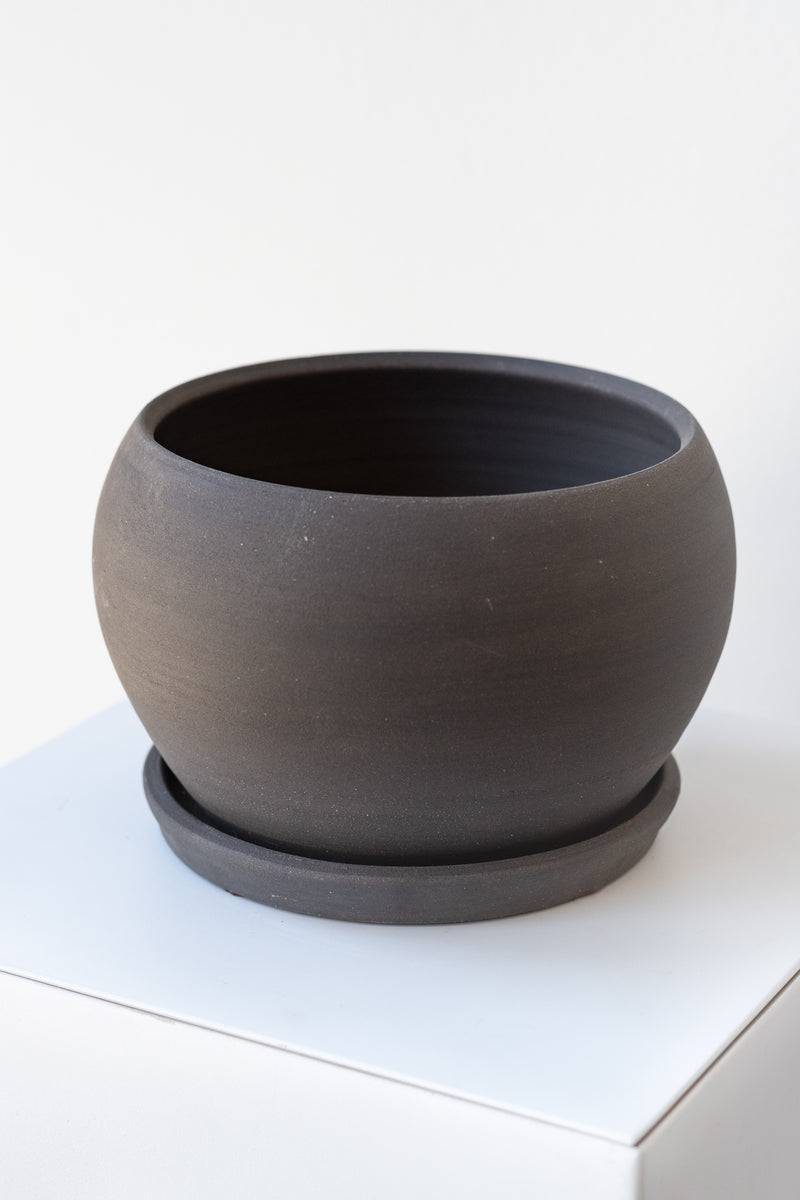 A round grey ceramic planter sits on a white surface in a white room. The planter is bubble-shaped and sits on a round drainage tray. The planter is empty. It is photographed closer and at an angle.