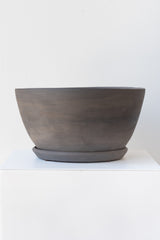 A round grey ceramic planter sits on a white surface in a white room. The planter is tapered, getting wider at the top, and it sits on a round drainage tray. The planter is empty. It is photographed straight on.