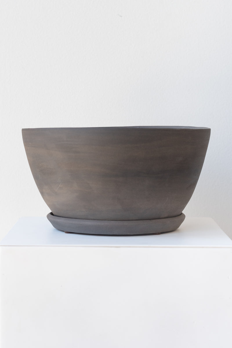 A round grey ceramic planter sits on a white surface in a white room. The planter is tapered, getting wider at the top, and it sits on a round drainage tray. The planter is empty. It is photographed straight on.