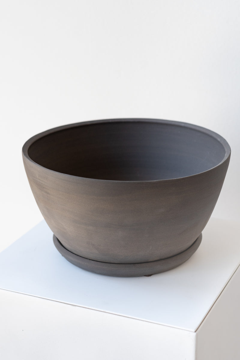 A round grey ceramic planter sits on a white surface in a white room. The planter is tapered, getting wider at the top, and it sits on a round drainage tray. The planter is empty. It is photographed closer and at an angle.