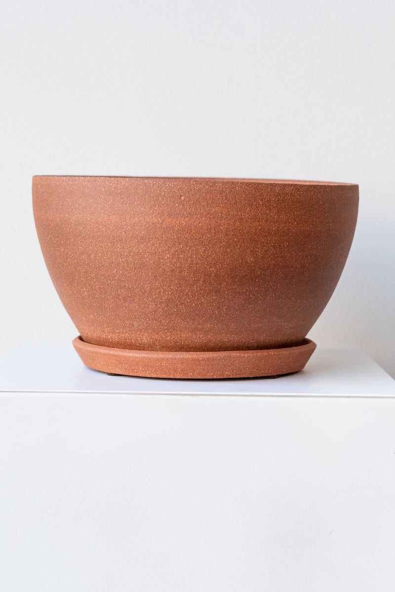 A round brick-colored ceramic planter sits on a white surface in a white room. The planter is tapered, getting wider at the top, and it sits on a round drainage tray. The planter is empty. It is photographed straight on.