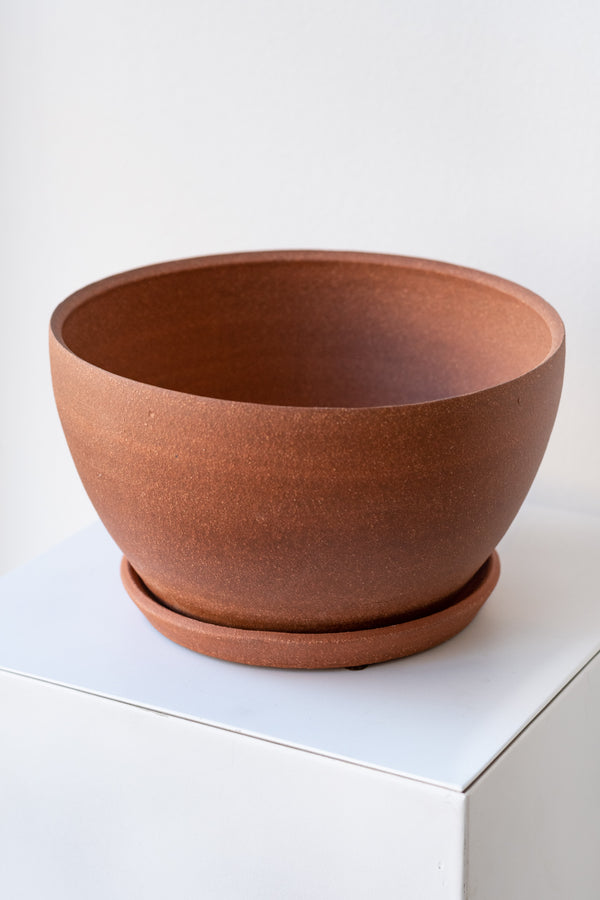 A round brick-colored ceramic planter sits on a white surface in a white room. The planter is tapered, getting wider at the top, and it sits on a round drainage tray. The planter is empty. It is photographed closer and at an angle.