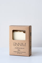 A bar of white soap sits in a brown paper box on a white surface in a white room. The box says "EINNIM, a lifestyle brand, natural products handmade in Chicago"