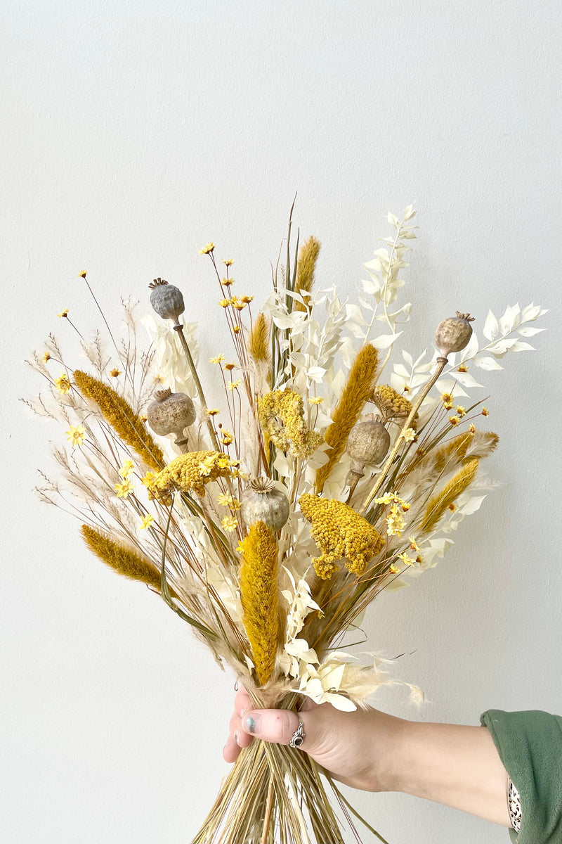 Yellow, white, and tan dried floral arrangement held by hand with ring and green sleeve. In front of white wall.