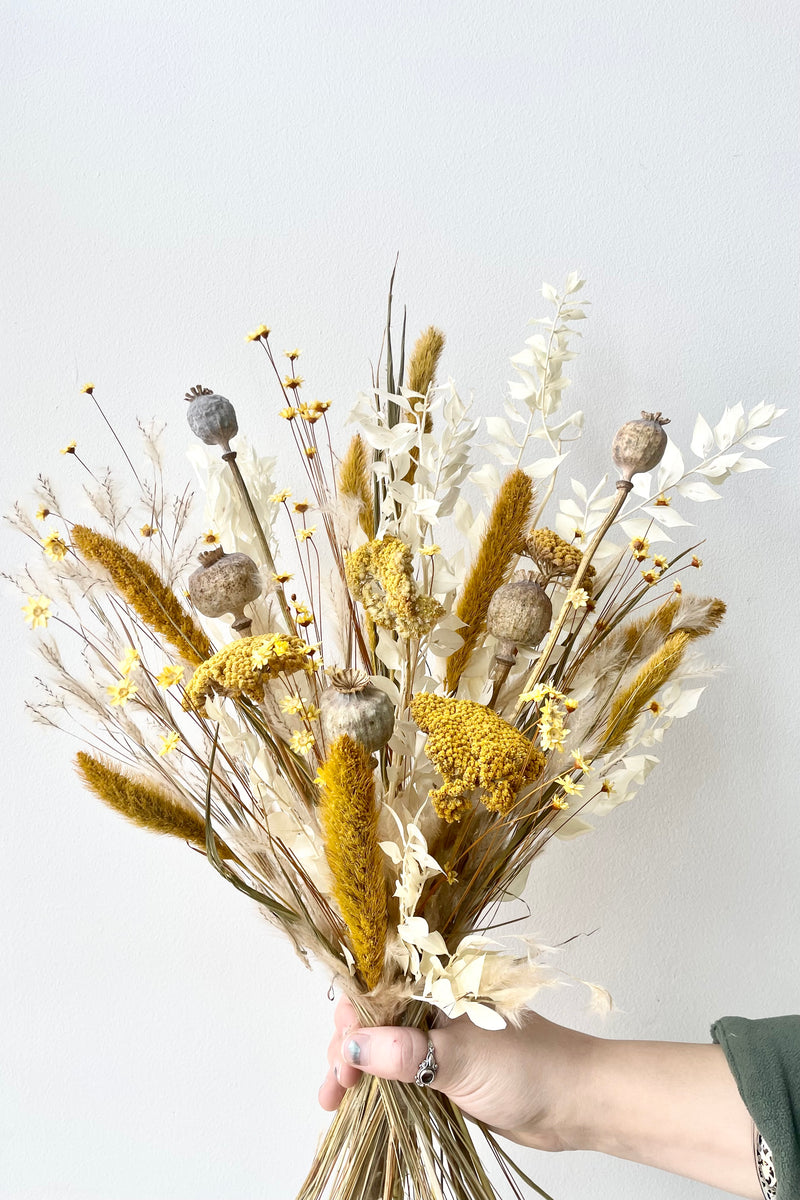 Dried floral arrangement with yellows, whites, and tans. Held by hand with rings and green shirt. White background.