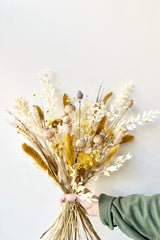 Hand holding dried floral with the colors yellow, white and tans in front of white background.