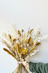 Lush dried floral arrangement with tans, whites, and yellows held by hand with green sleeve. In front of white background.